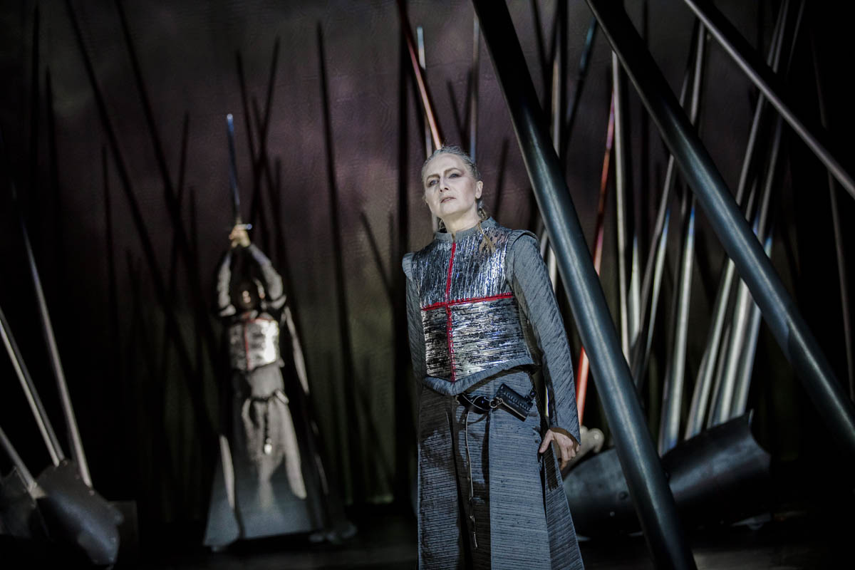 Kobieta ubrana w zbroję rycerską stoi na tle ustawionych nieregularnie wysokich dragów imitujących kopie rycerskie. W tle mężczyzna w zbroi rycerskiej, który w uniesionych dłoniach trzyma miecz.