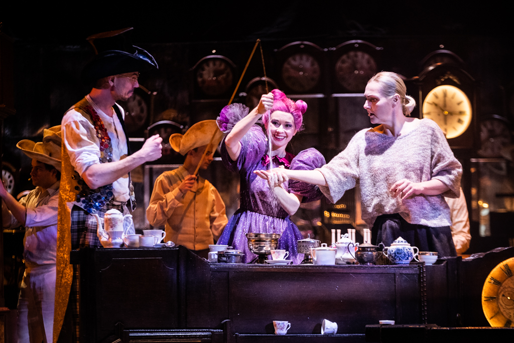 Alicja, wraz ze swoimi towarzyszami: kobietą w różowej peruce i z mężczyzną w czarnym kapeluszu bawi się czasem. Przesypują piasek z klepsydry. W tle widać zegary, niektóre z nich są podświetlone na pomarańczowo.