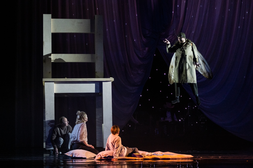 Jest gwiaździsta noc. Przy wielkim, białym krześle siedzi Alicja wraz z mężczyzną i z kobietą. Po prawej stronie unosi się mężczyzna, który ma z tyłu przyczepione skrzydła ćmy.