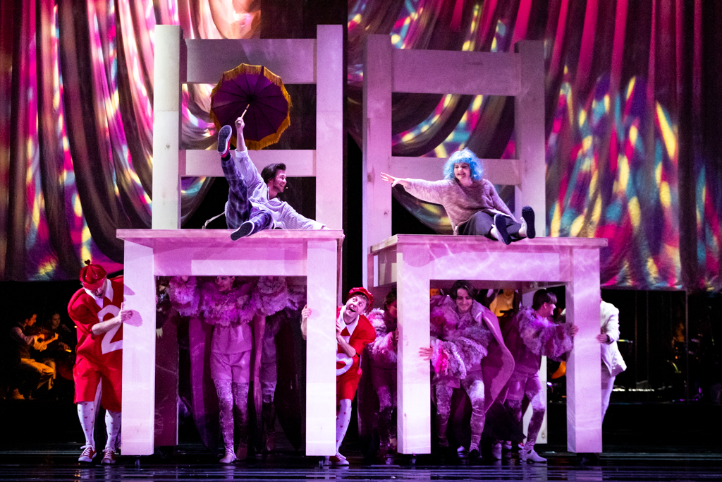 Na wielkich, fioletowych krzesłach siedzą Alicja oraz mężczyzna trzymający w prawej ręce rozłożoną parasolkę. Alicja ma na głowie niebieską perukę i wskazuje dłonią na swojego towarzysza. Pod krzesłami są aktorzy ubrani w fioletowe kostiumy.