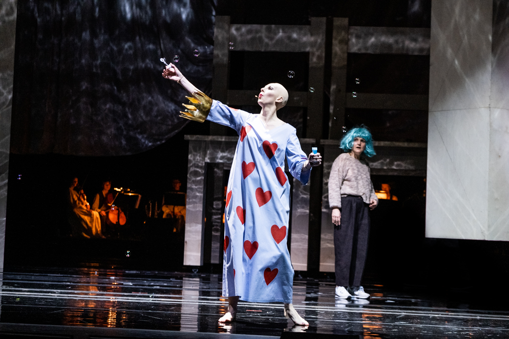 Królowa Kier, ubrana w długą białą sukienkę w czerwone serca, trzyma w ręku klucz. Z boku przygląda się jej Alicja - w sportowym stroju i w niebieskiej peruce. Z tyłu sceny widać grających muzyków.