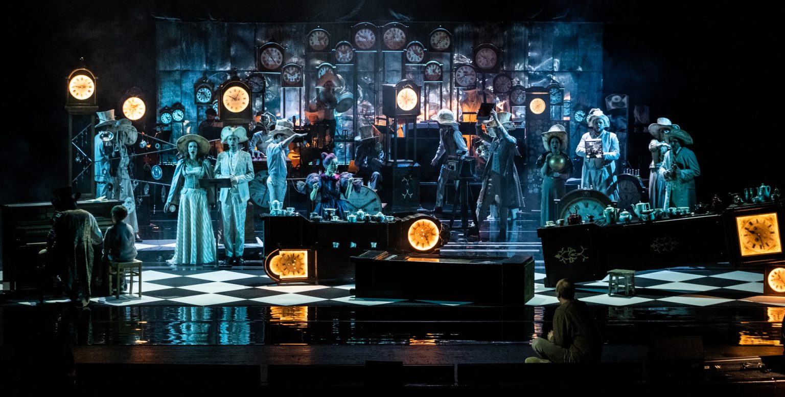 Na zdjęciu widać Cmentarzysko zegarów. Mężczyźni i kobiety w kapeluszach trzymają różne przedmioty w rękach. Wszędzie są widoczne zegary, niektóre z nich stoją, wiszą, są podświetlone na pomarańczowo, leżą na podłodze w biało - czarną kratę, przypominającą szachownicę.