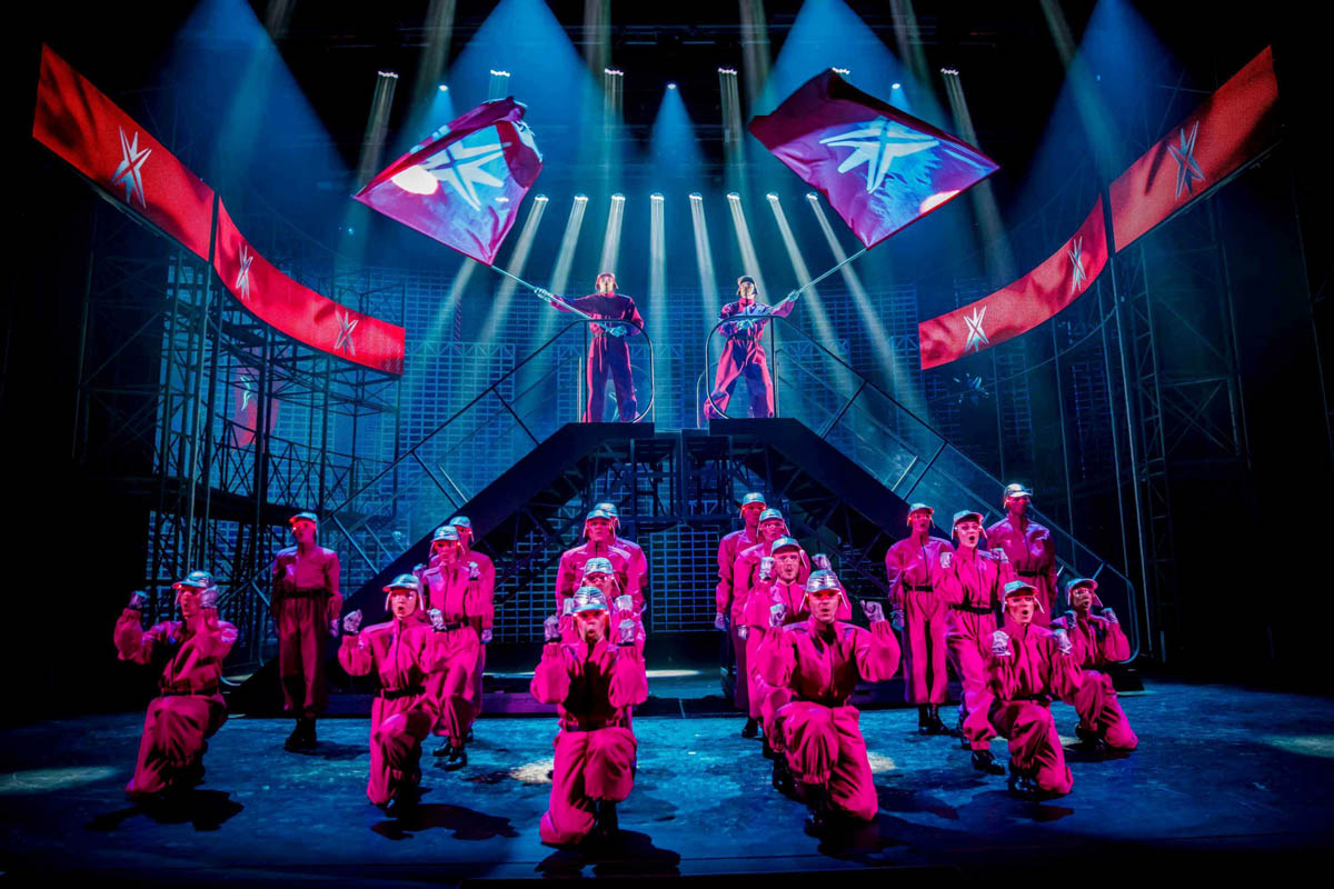 Klęcząca grupa kilkunastu postaci ubranych w jednakowe kombinezony nawiązujące do stylu odzieży ochronnej i w jednakowych błyszczących czapkach sportowych na głowach. W tle dwie z nich stoją na wysokich podestach, a w dłoniach trzymają powiewające sztandary.