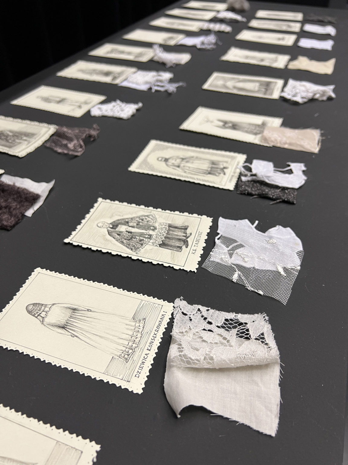 Na pierwszym planie znajduje się kawałek białej koronki, a w tle kilka innych tkanin. Obok rozłożone są obrazki przedstawiające kobiety w strojach tradycyjnych