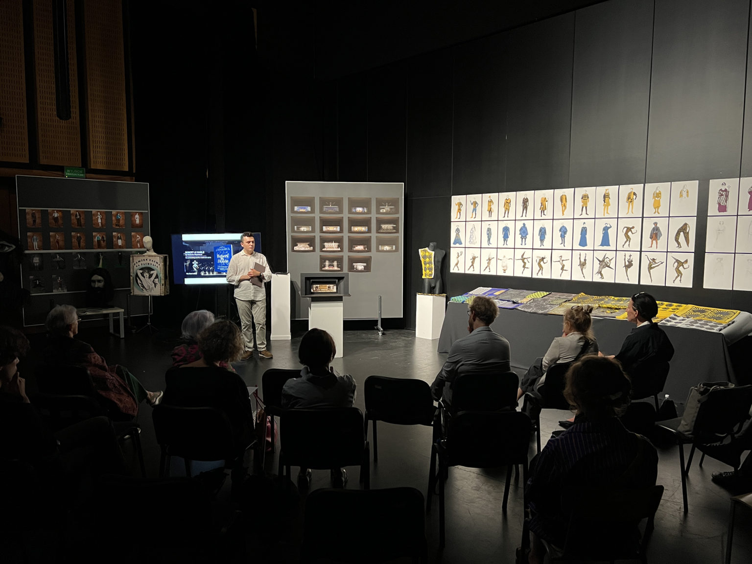 Mężczyzna stoi pomiędzy dwoma manekinami przed grupą ludzi, wszyscy siedzą na krzesłach i są zwróceni w jego stronę. Za nim na ścianie wiszą obrazy,