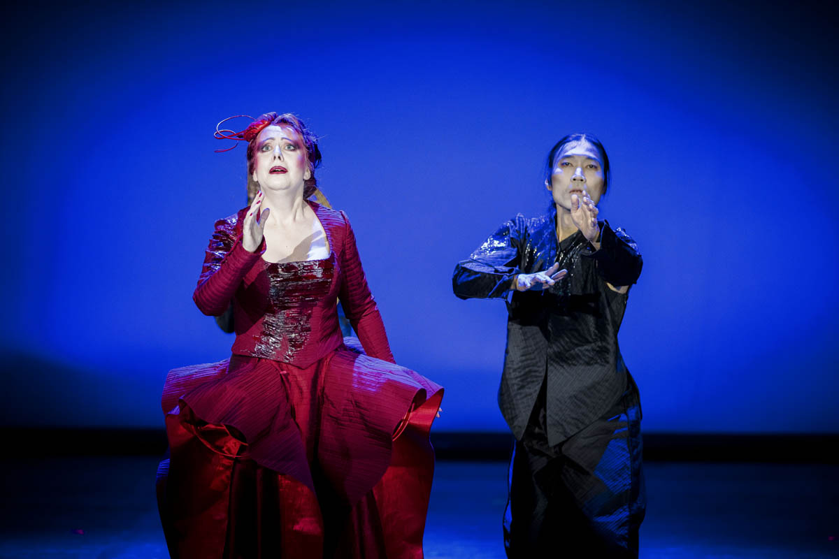 Kobieta w rozbudowanej, długiej sukni stoi obok mężczyzny w ciemnym garniturze. Oboje z wyciągniętymi do przodu ramionami.