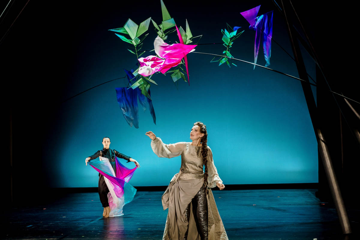 Kobieta w długiej sukni podnosi dłoń w kierunku kobiety w obcisłym stroju, trzymającej w dłoniach powiewające kawałki cienkiej tkaniny. Nad ich głowami powiewają nieregularnie rozmieszczone cienkie tkaniny.