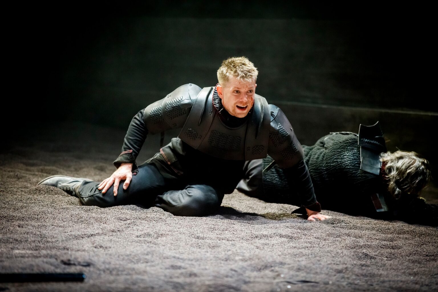 Aktor ubrany w skórzaną zbroję, leżący na scenie pokrytej ciemną ziemią