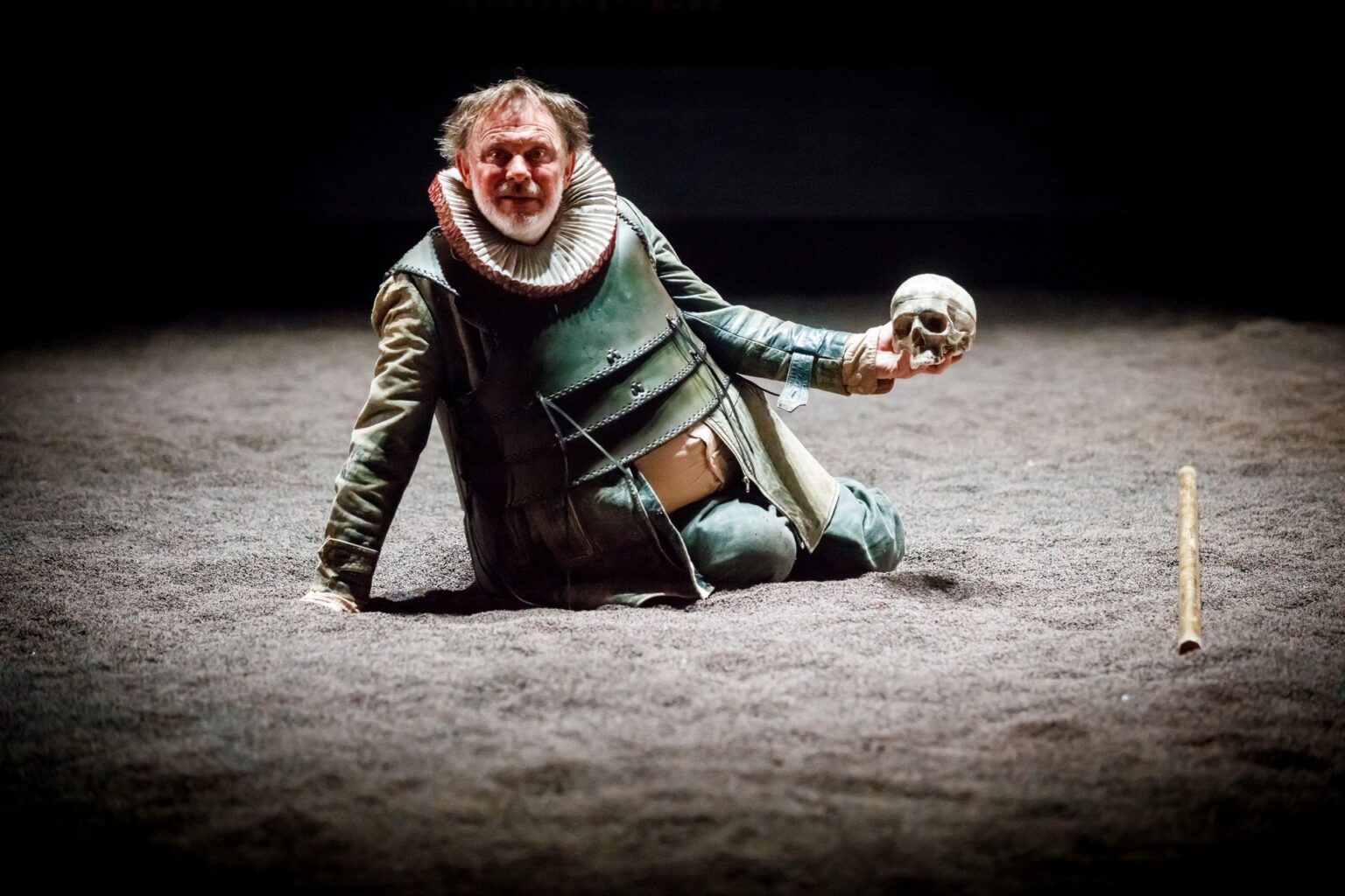Aktor ubrany w skórzaną zbroję, leżący na scenie pokrytej ciemną ziemią