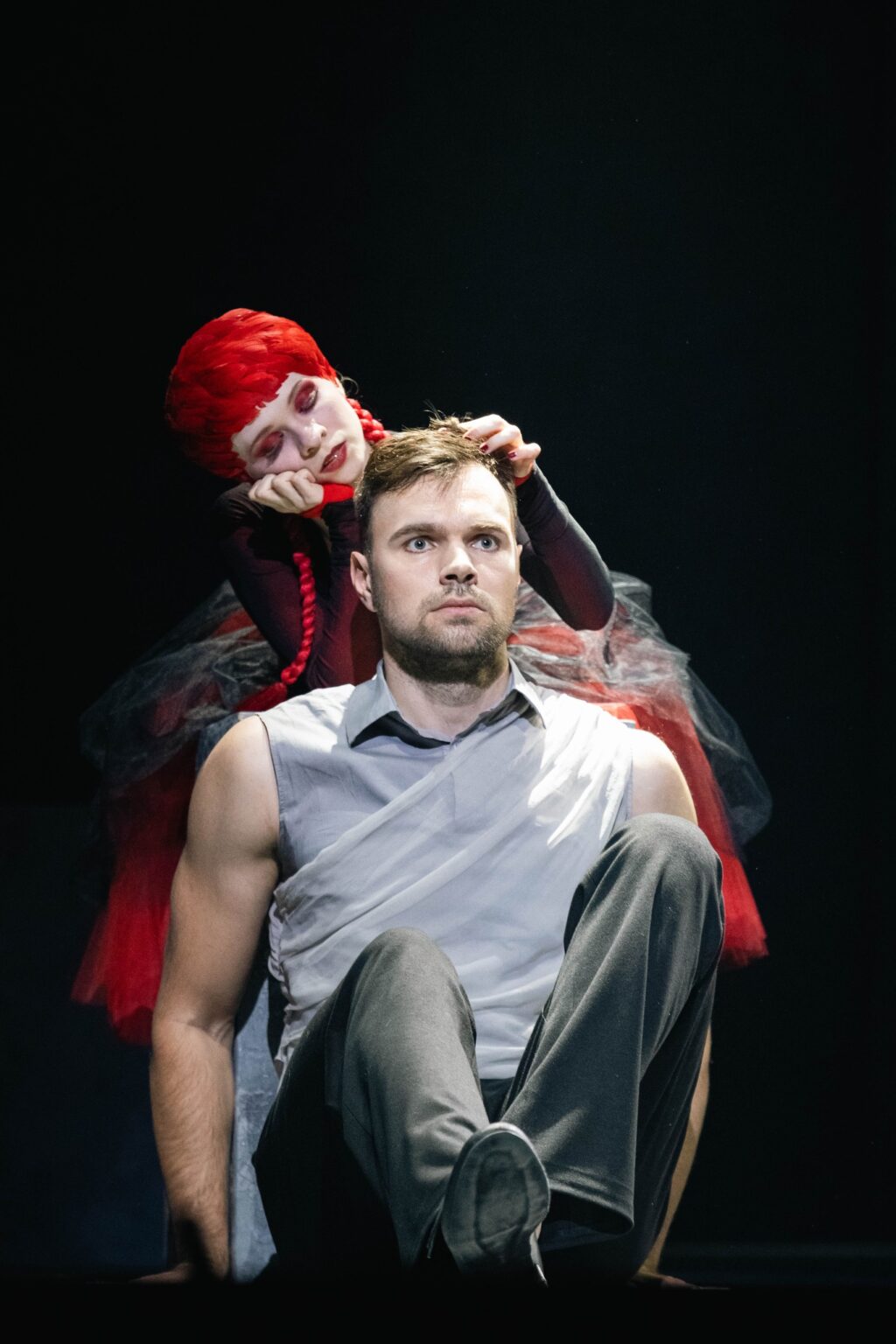 Kobieta w czerwonej peruce wykonanej z piór, siedzi za plecami przerażonego mężczyzny