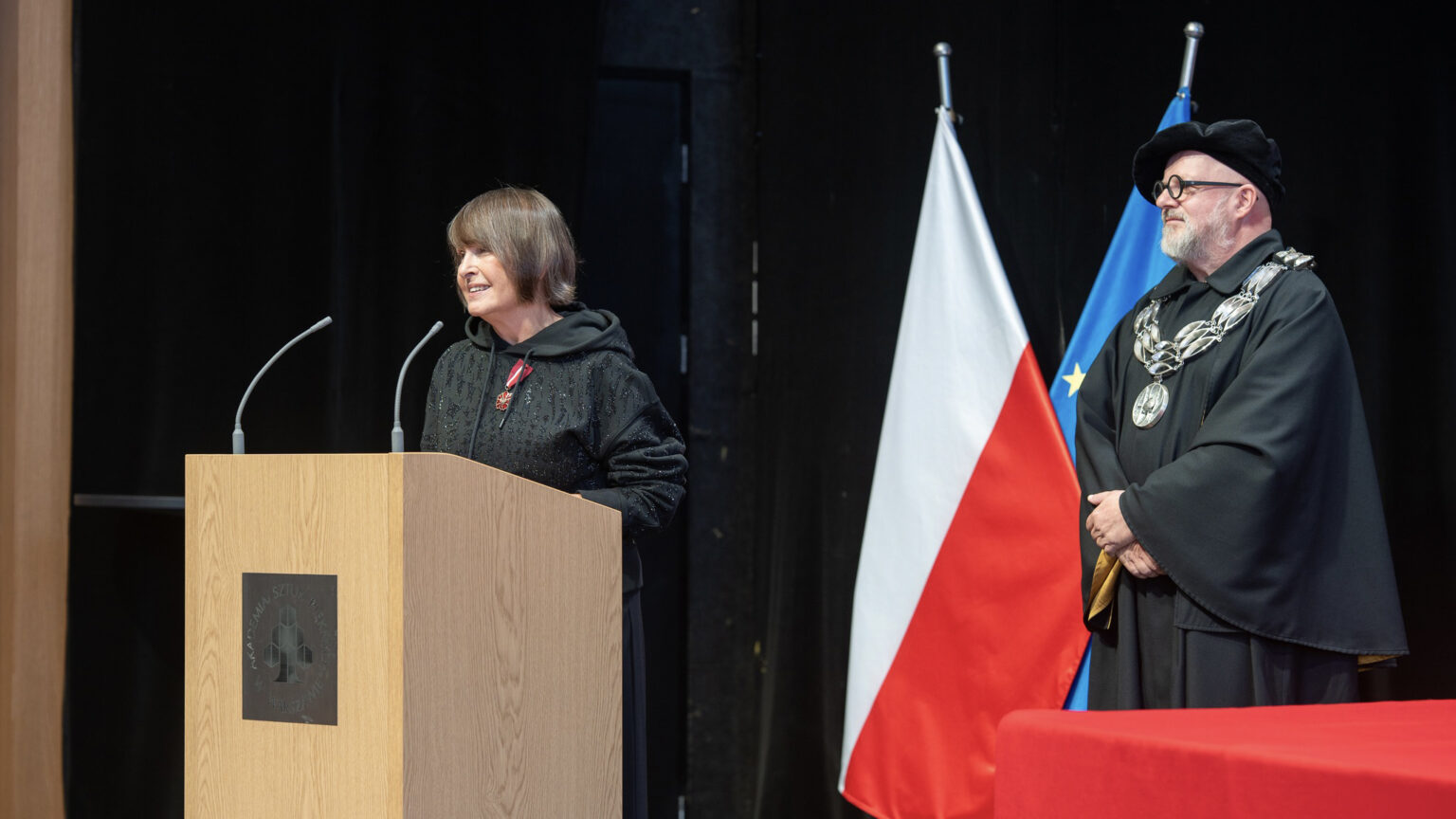 Na zdjęciu kobieta stojąca przy mównicy, z jej lewej strony stoi mężczyzna odświętnie ubrany. Zdjęcie przedstawia moment wręczenia Brązowego Medalu "Zasłużony Kulturze Gloria Artis"