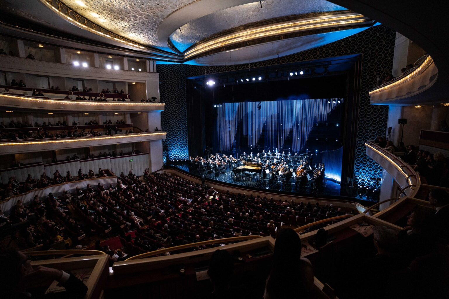 Zdjęcie przedstawia widownię i scenę Teatru Wielkiego – Opery Narodowej w Warszawie. Na scenie znajduje się całą orkiestra na tle pięknej, minimalistycznej scenografii.