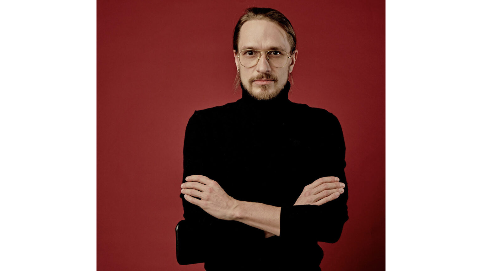 Zdjęcie Mateusza Atmana, na czerwonym tle. Mateusz ubrany jest w czarny golf, ręce założone ma na piersi.