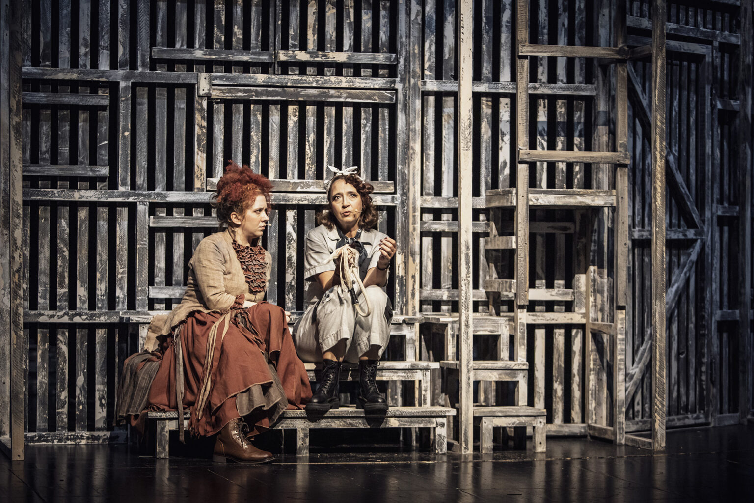 Dwie kobiety siedzące na małych schodach. Jedna z nich ubrana jest w kombinezon z dużą ilością kieszeni. Druga natomiast w spódnicę oraz żakiet.