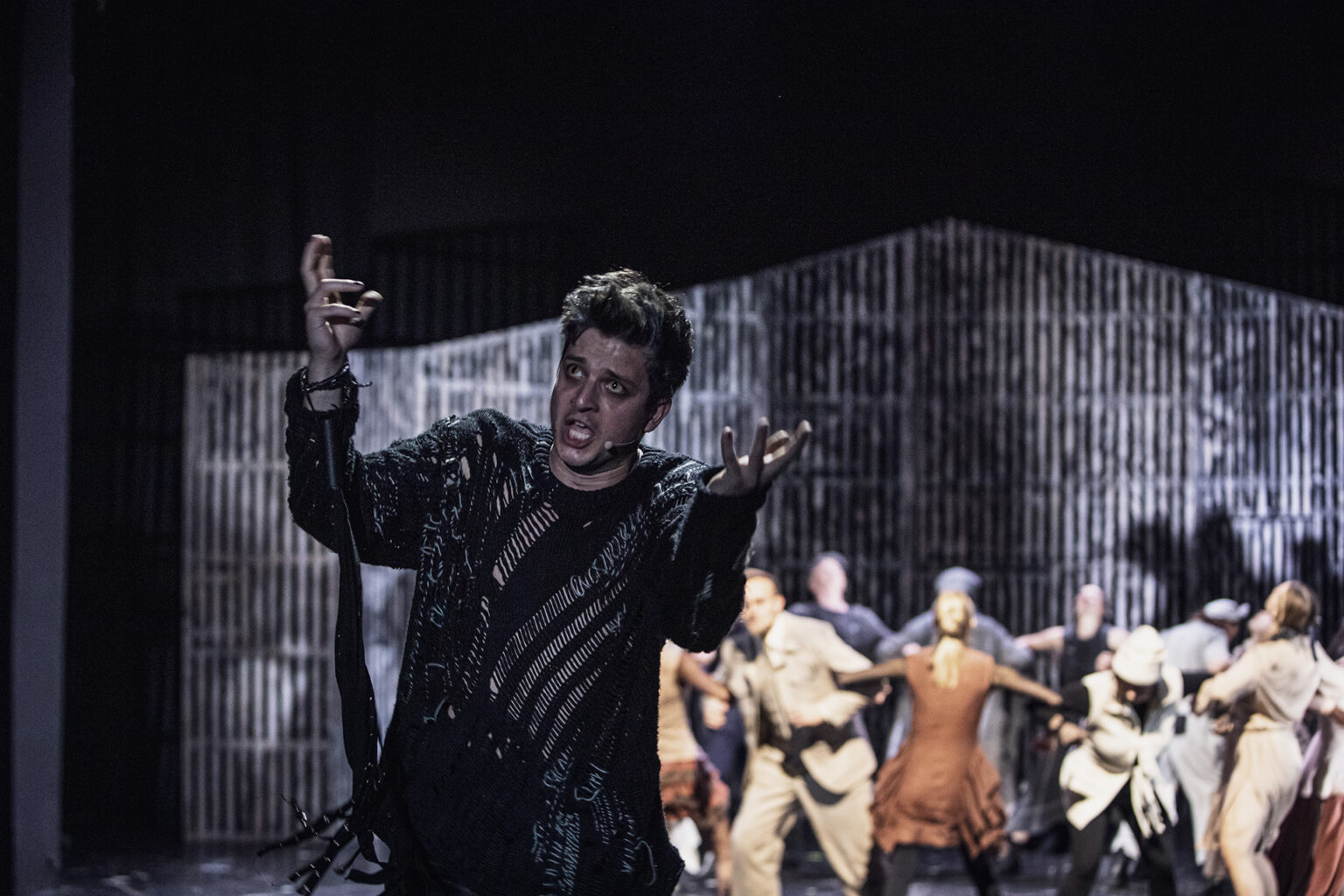 Zdjęcie mężczyzny w dziurawym swetrze, stojącego na tle tańczącej grupy.