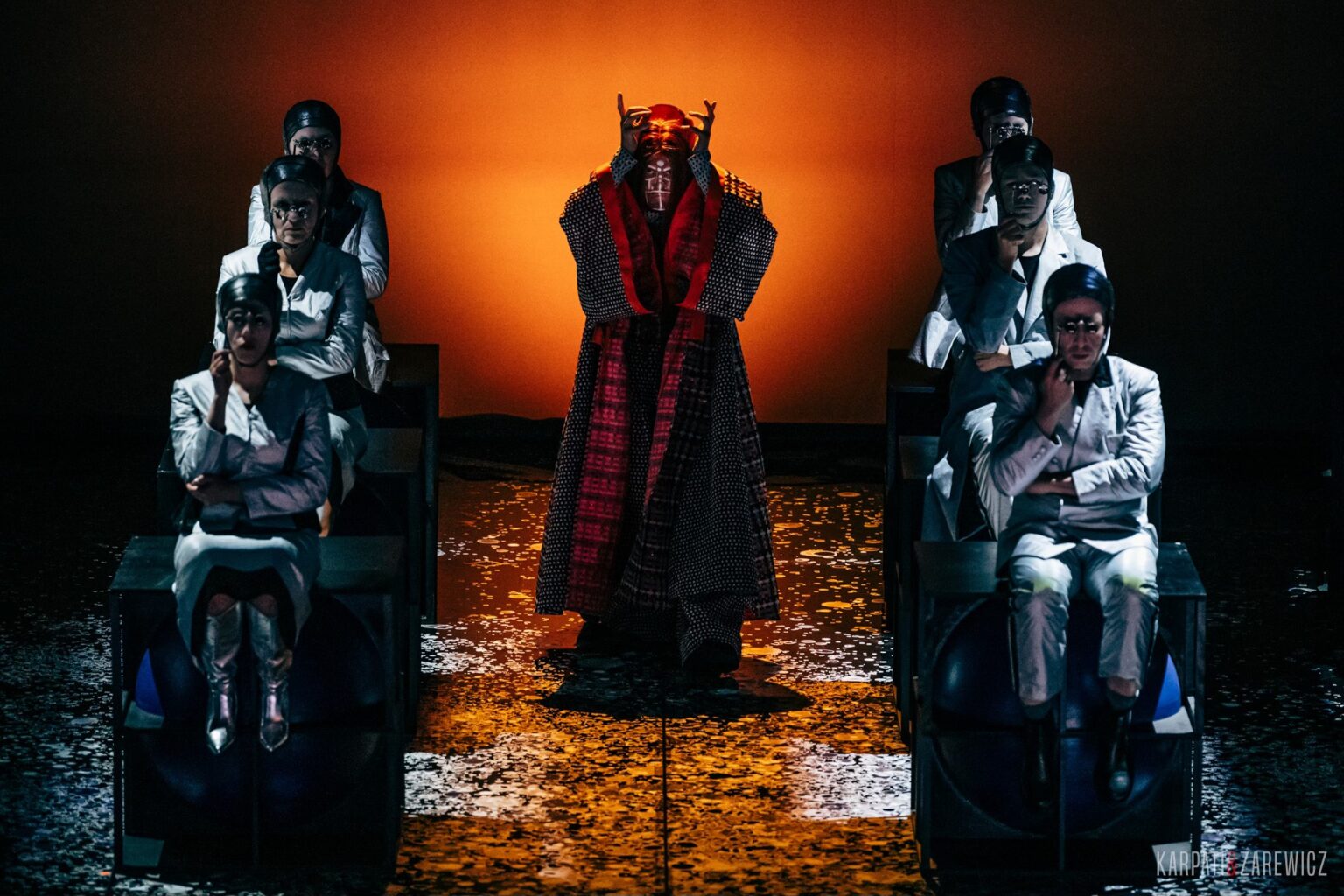 Zdjęcie przedstawia grupę ludzi ubranych w identyczne szare kostiumy, siedzących na stołkach, po środku której stoi postać ubrana w długi czerwony płaszcz.