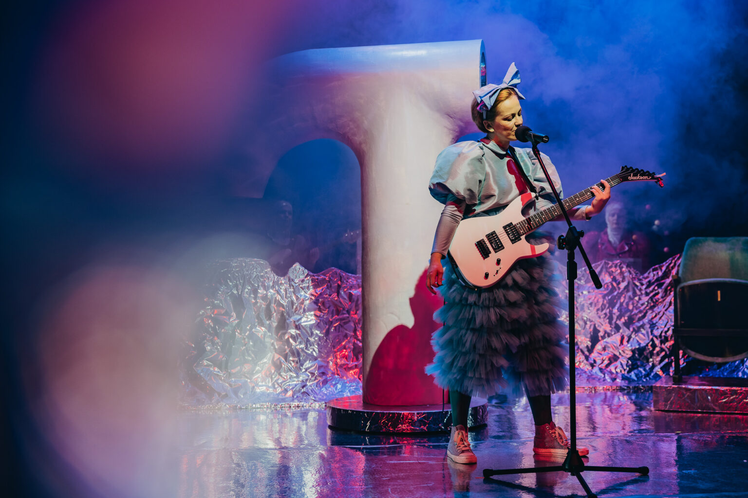 Kobieta stojąca przy mikrofonie, ubrana w falbaniastą sukienkę. Kobieta trzyma w ręku gitarę.