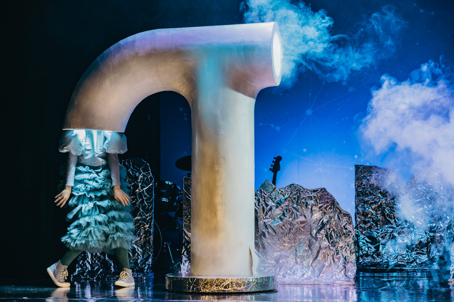 Na zdjęciu stoi kobieta, której głowa schowana jest w gigantycznej rurze, przypominającej komin.