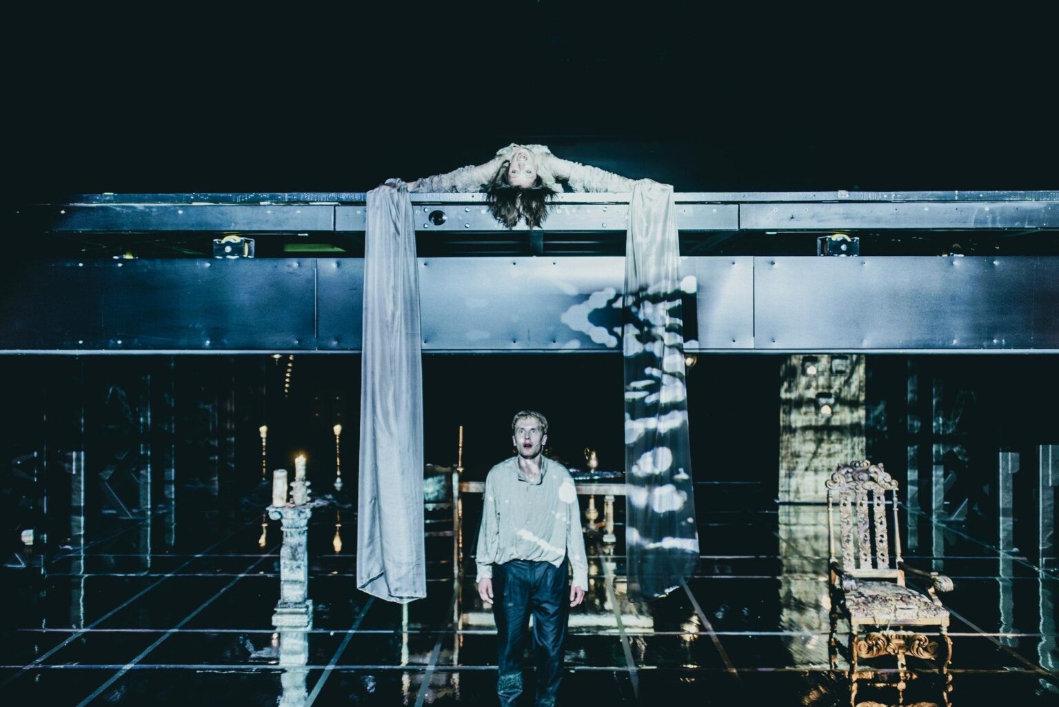 Scena przedstawia dwupoziomową scenografię. Na górnym piętrze leży tajemnicza postać z rąk której zwisa biała tkanina. Pod nią stoi przerażony mężczyzna. Obok niego świecznik i krzesło.