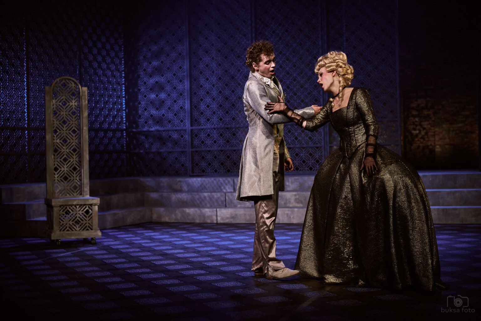 Zdjęcie przedstawiające romantyczną scenę dwóch osób: młodej kobiety ubranej w złotą suknię oraz młodego mężczyzny w jasnym kostiumie.