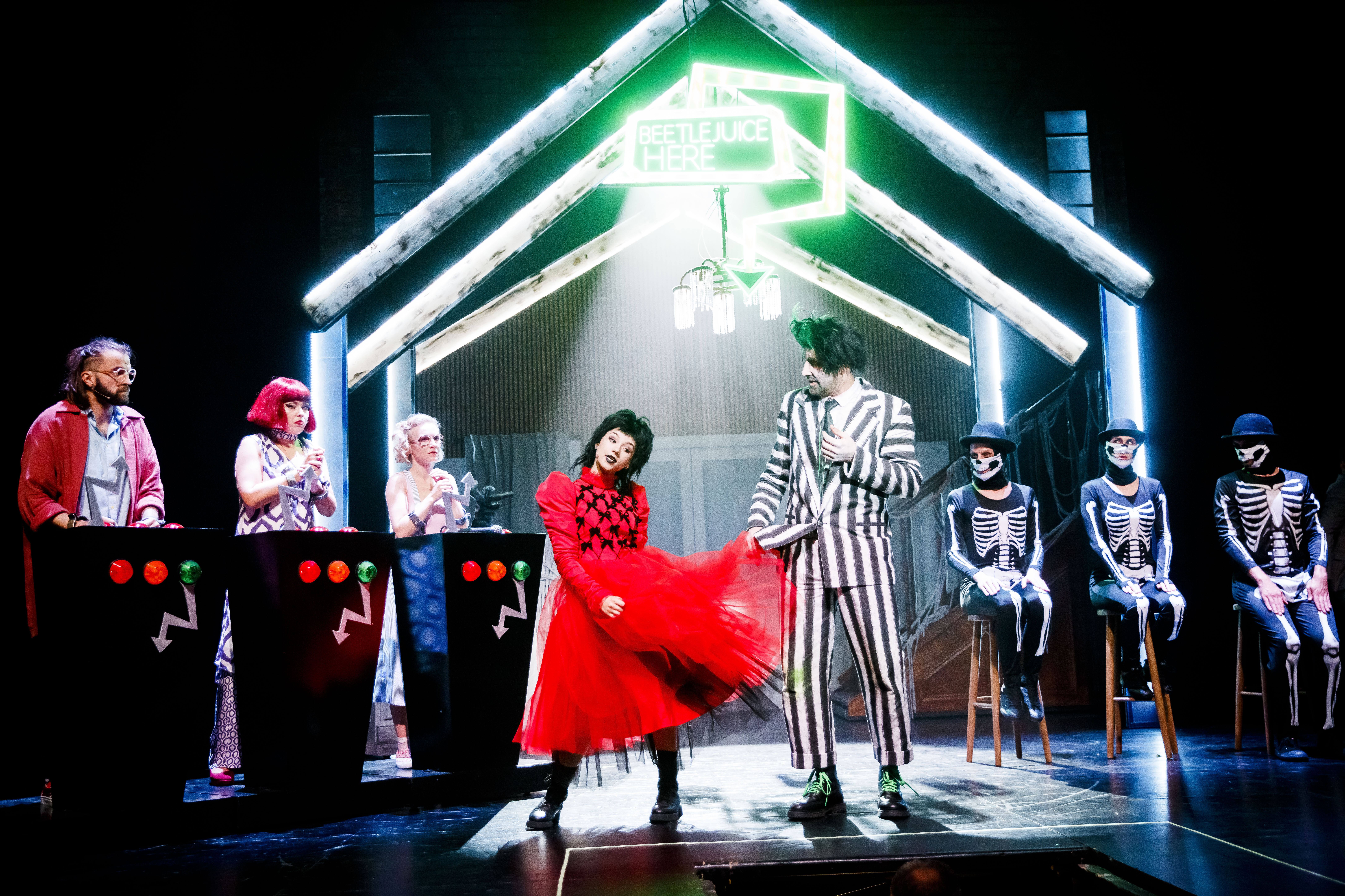 Zdjęcie przedstawiające dziewczynę w czarnej peruce, w czerwonej sukience, mężczyznę w garniturze w paski, kobietę w czerwonej peruce, blond kobietę w okularach oraz dwie osoby przebrane za trupy.