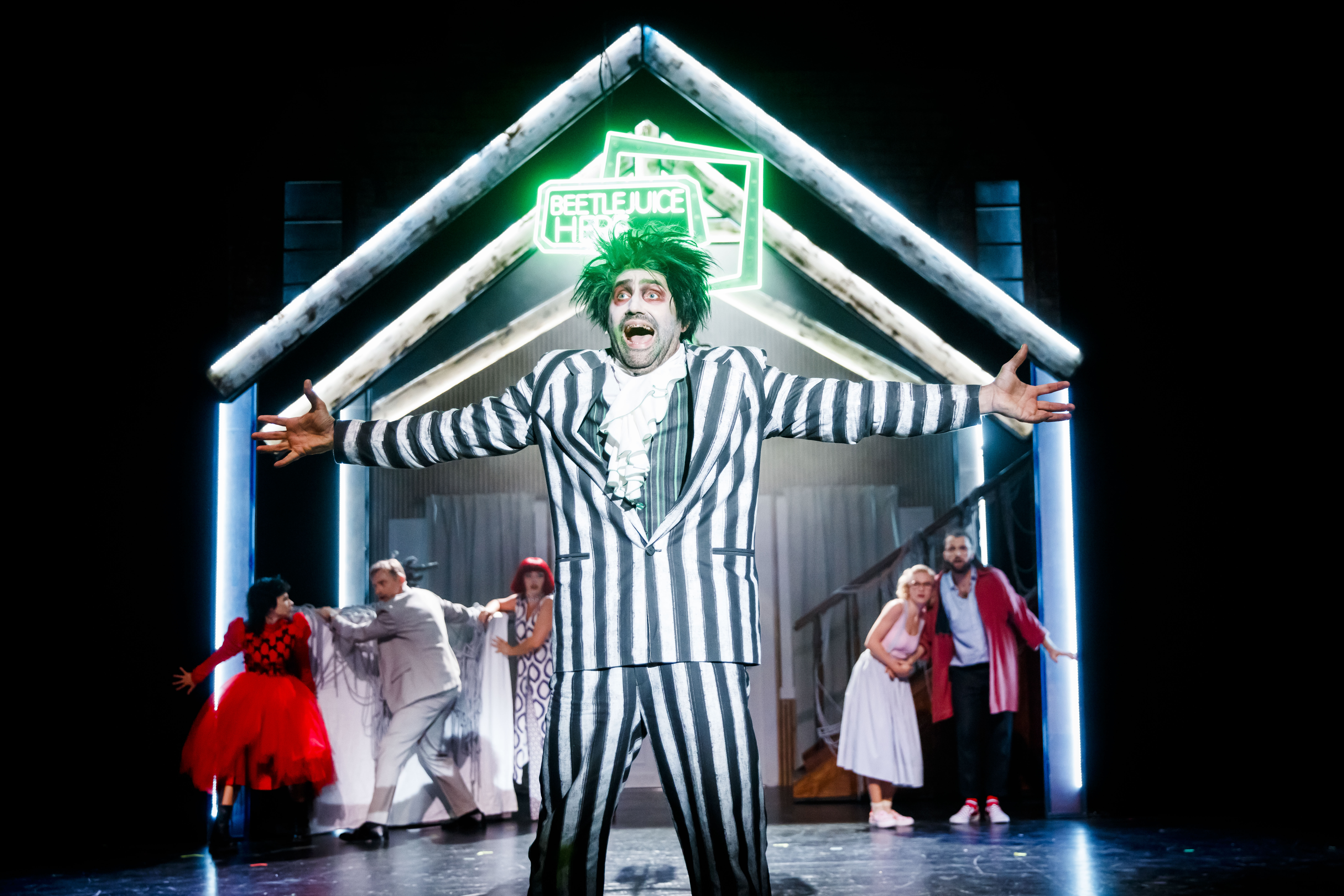 Zdjęcie grupowej sceny, na czele której stoi rozemocjonowany mężczyzna ubrany w garnitur w paski oraz czarno-zieloną perukę.