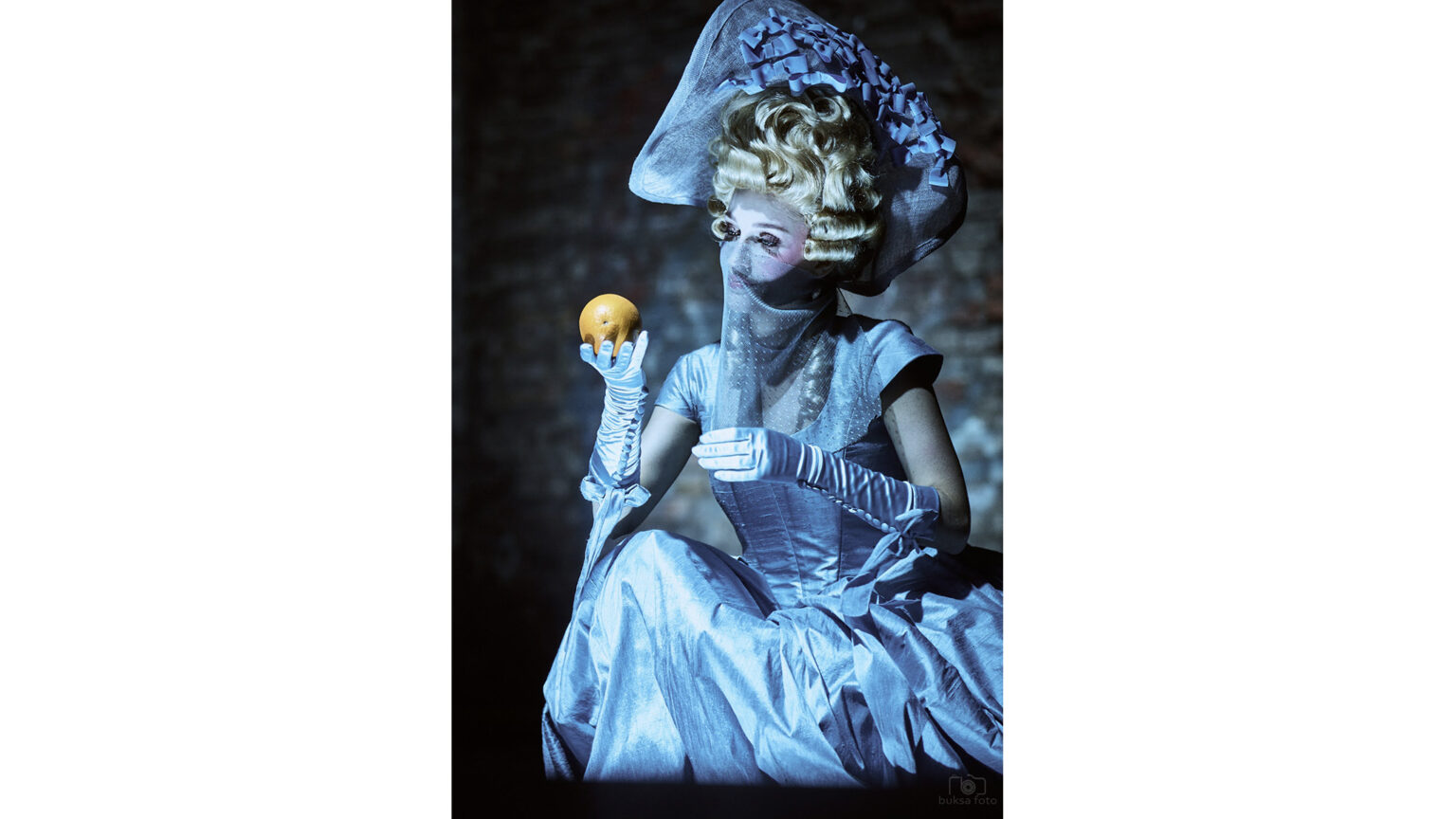 Zdjęcie młodej kobiety, w niebieskiej sukience, trzymającej pomarańczę w ręku.