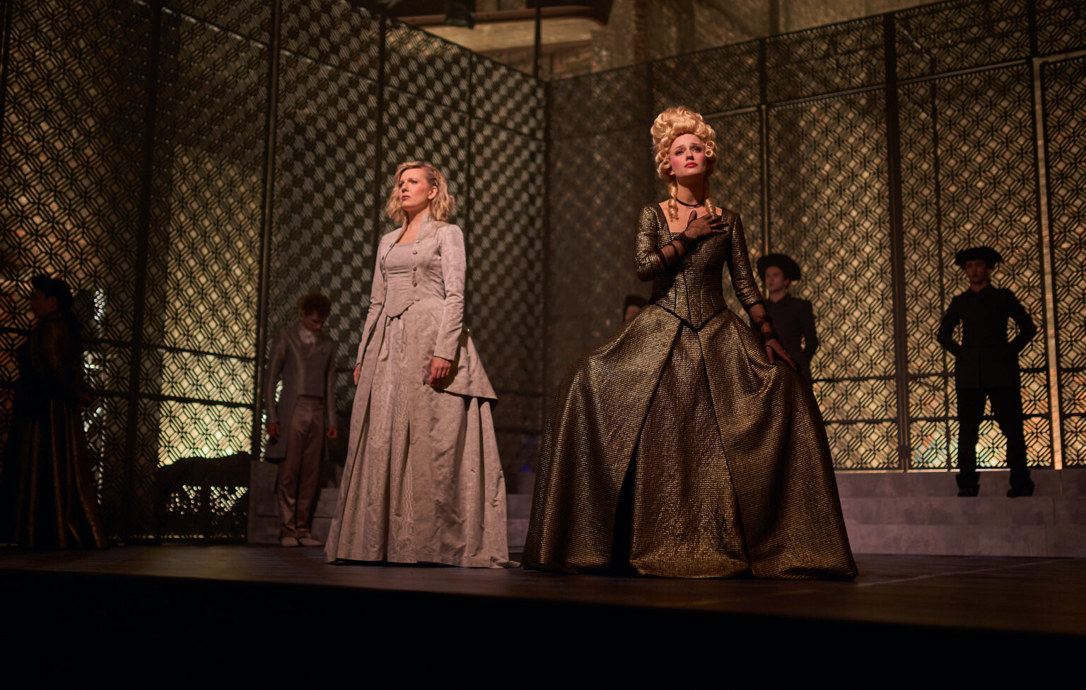 Dwie kobiety ubrane w kostiumy inspirowane modą rokokową, stojące na tle ażurowej scenografii.