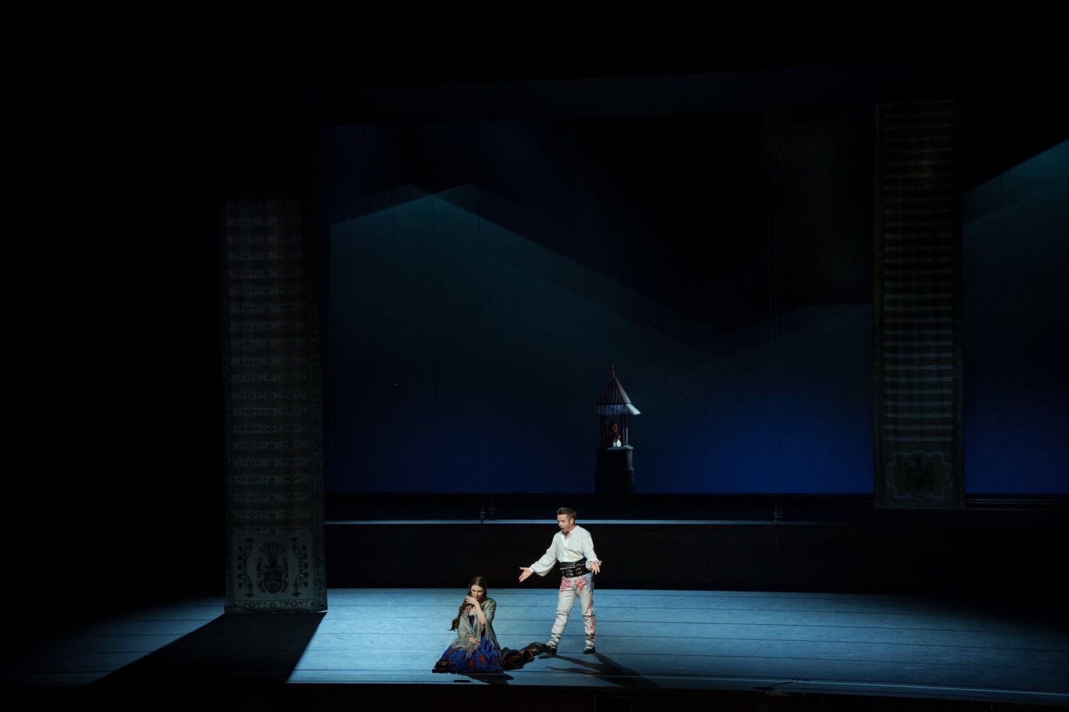 Zdjęcie przedstawia śpiewaka ubranego w strój góralski. Mężczyzna stoi przy kobiecie siedzącej na ziemi.