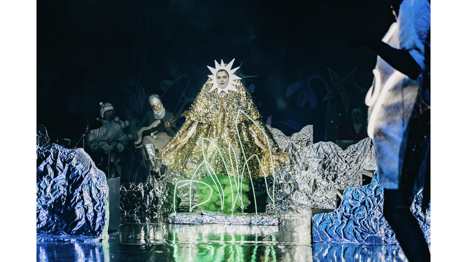 Kobieta - słońce, ubrana w złoty kostium, siedzi na tle srebrnej scenografii.