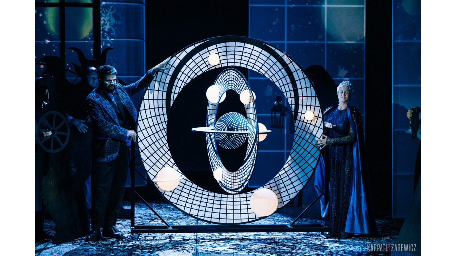 Zdjęcie przedstawia dwie postaci podtrzymujące ciekawy obiekt, przypominający swoim kształtem układ słoneczny.