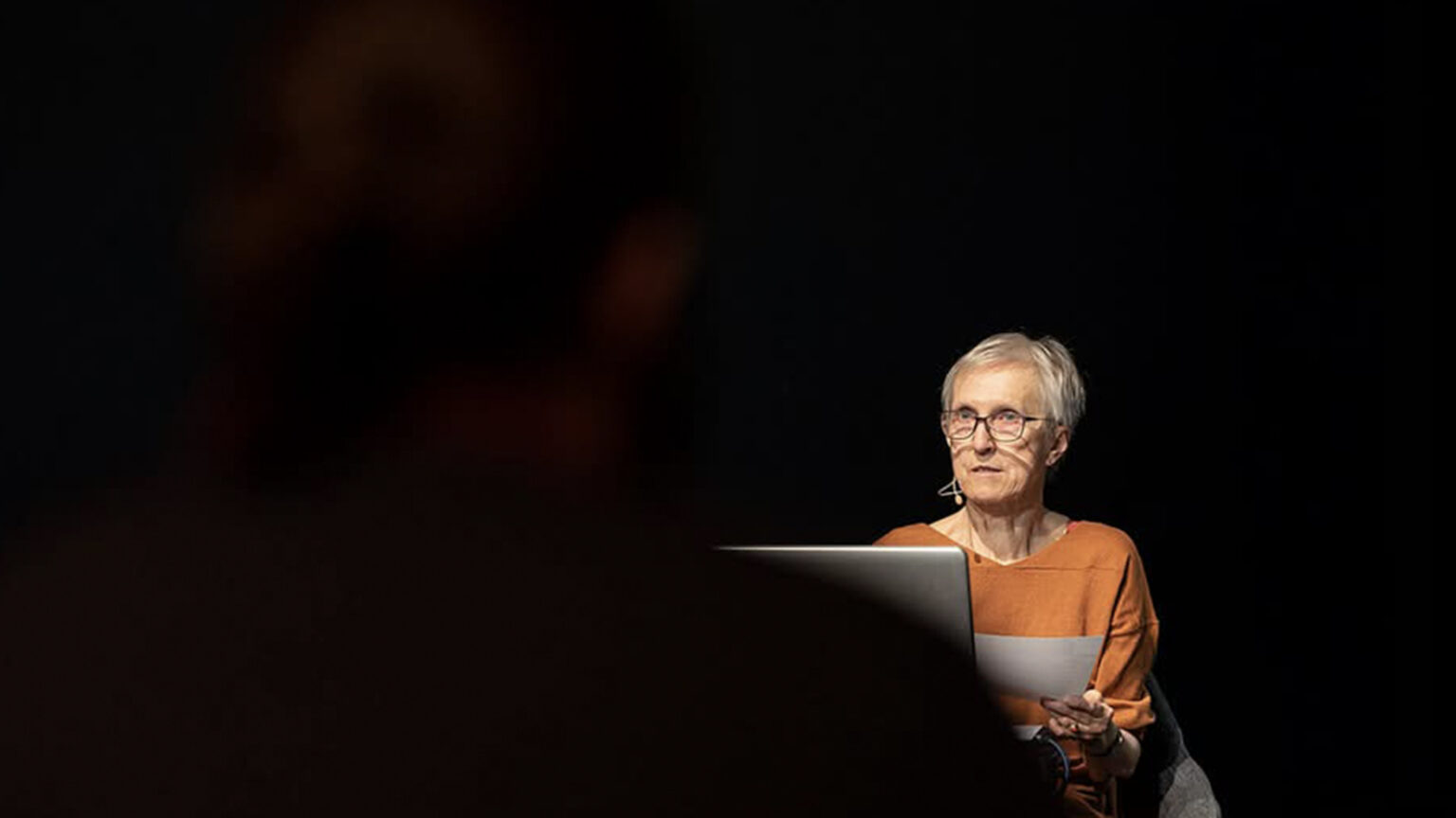 Kobieta opowiadając siedzi w krześle. MA krótkie włosy, okulary i pomarańczowy sweter.