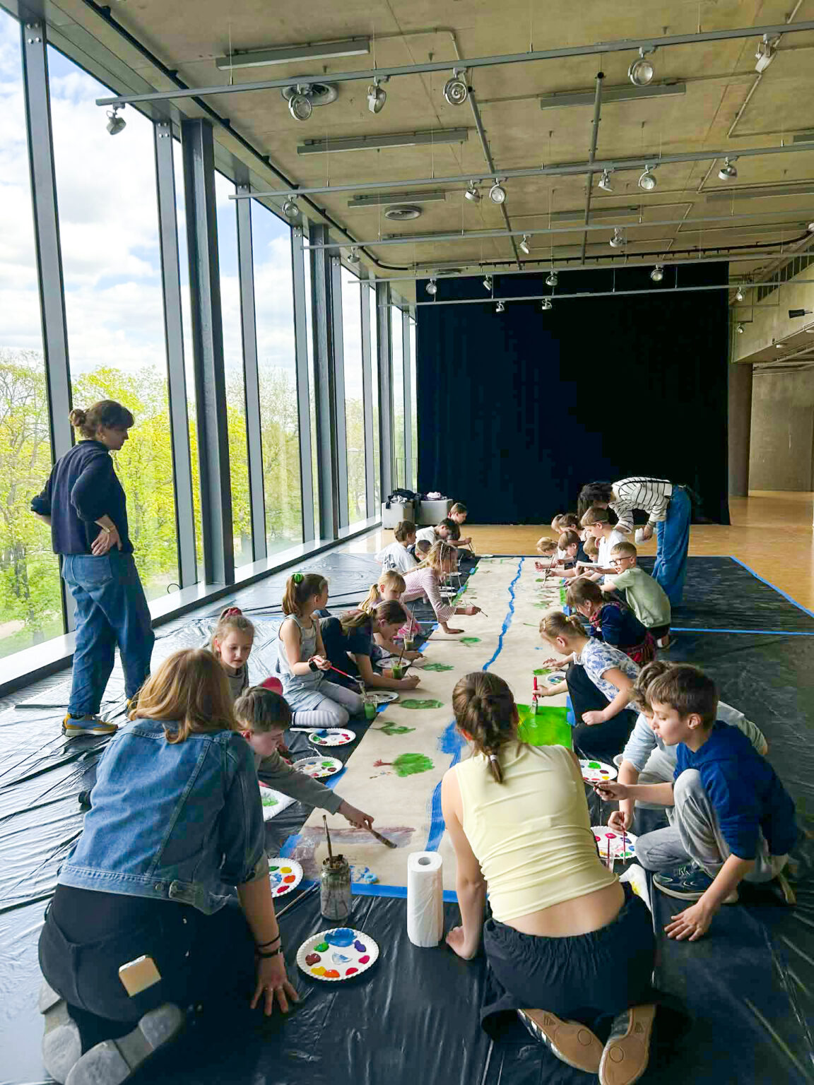 Grupa dzieci malująca długi pas szaro-brązowego papieru kolorowymi farbami. Na pierwszym planie klęczą dwie osoby dorosłe. Z lewej strony znajduje się duże okno z widokiem na drzewa na tle, którego stoi młoda kobieta.