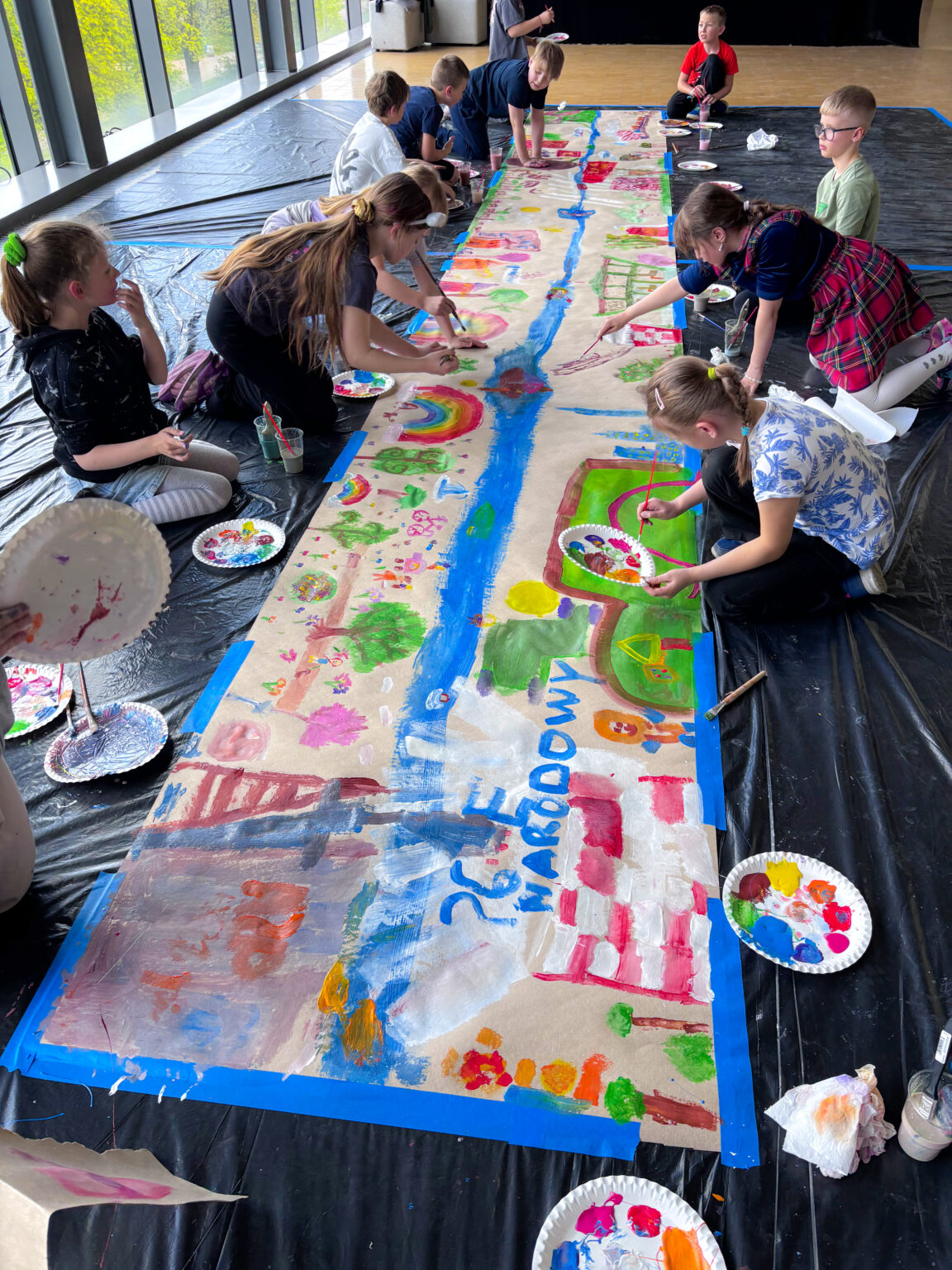 Grupa dzieci siedzi na podłodze wyłożonej czarną folią i maluje pędzlami i kolorowymi farbami długi pas szaro-brązowego papieru obklejonego niebieską taśmą.