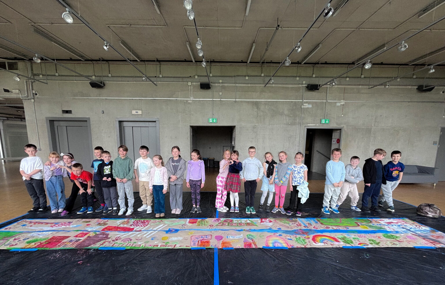 Grupa stojących dzieci pozujących do zdjęcia na tle szarej ściany. Przed nimi, na podłodze, leży długi pas szaro-brązowego papieru pomalowany kolorowymi farbami.