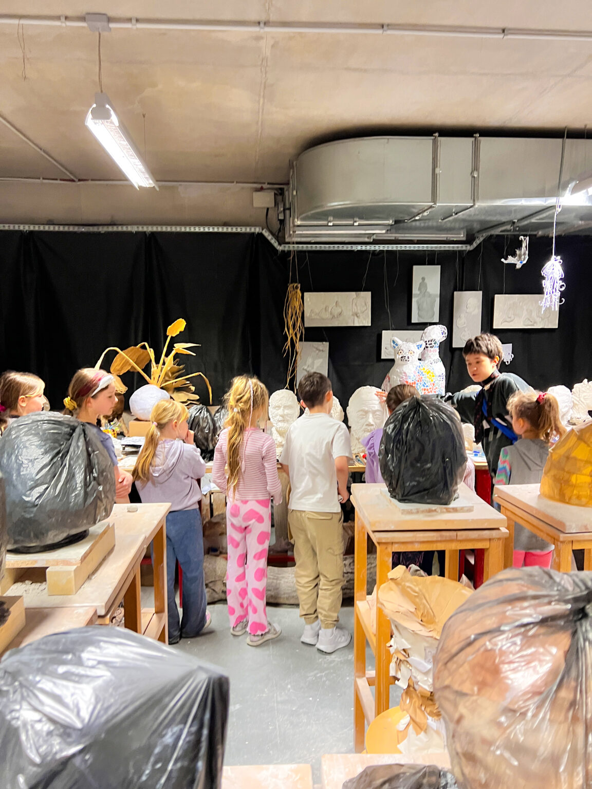 Osoba dorosła stoi z grupą dzieci w pomieszczeniu. Oglądają rzeźby postawione na stole. Na pierwszym planie stoją wysokie stojaki na których położone są zafoliowane workami na śmieci owalne formy.