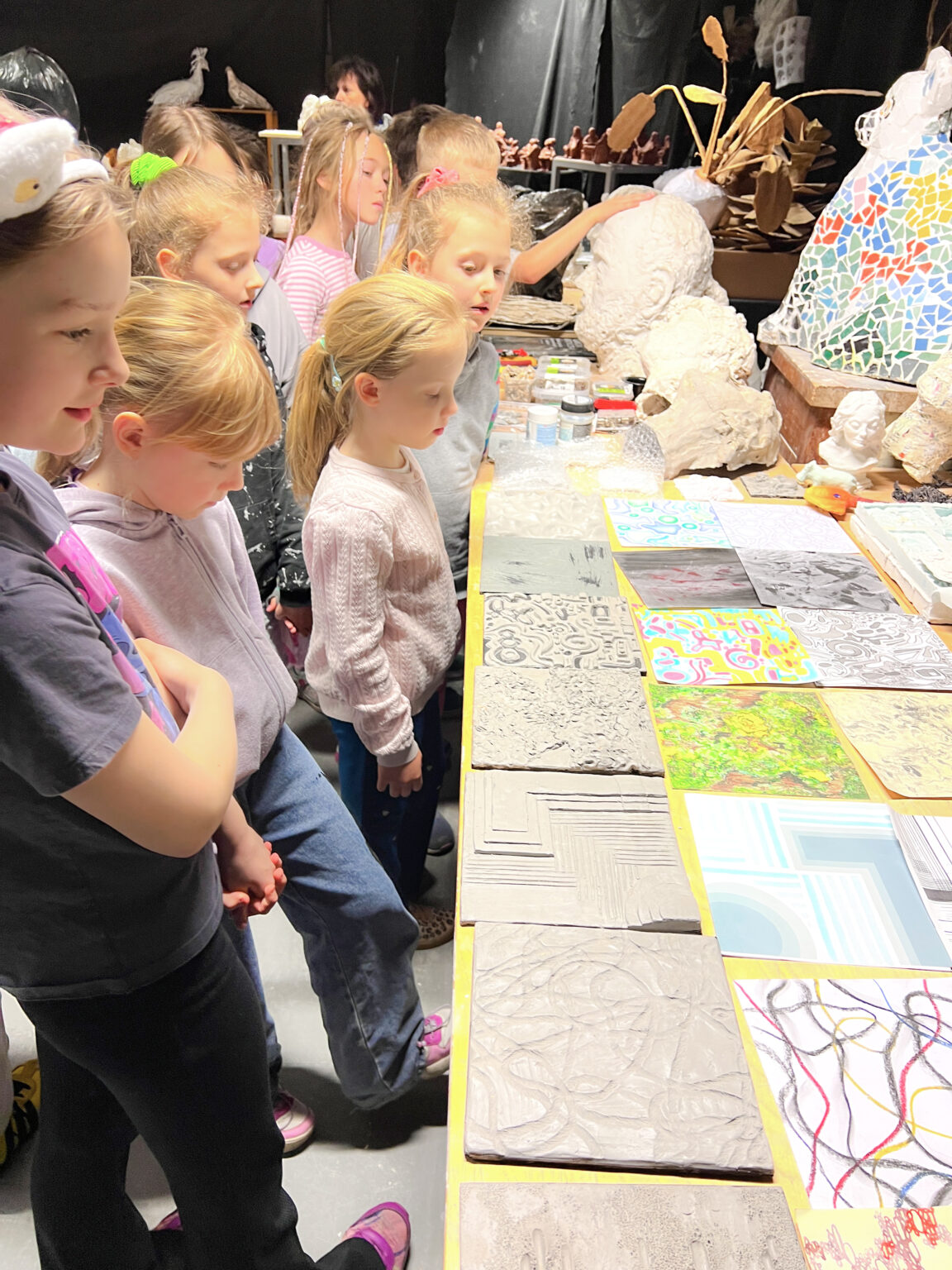 Z lewej strony znajduje się grupa dzieci i przygląda płaskim wzorzystym formom z gliny oraz kolorowym rysunkom ułożonym obok siebie na stole po prawej stronie.