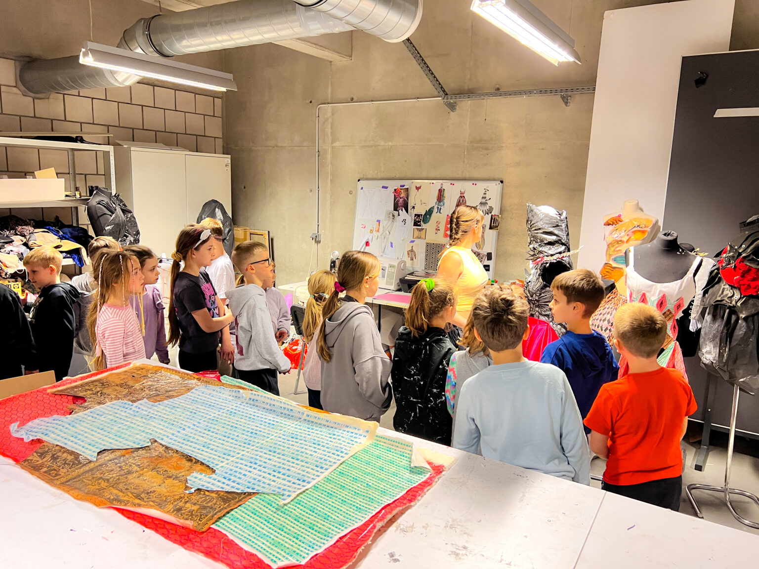 Osoba dorosła wraz z grupą dzieci stoi w pomieszczeniu i ogląda formy abstrakcyjnych kostiumów przymocowanych do czarnych materiałowych tułowi manekinów krawieckich. Na pierwszym planie jest biały stół, a na nim znajdują się wzorzyste kolorowe płaszczyzny.