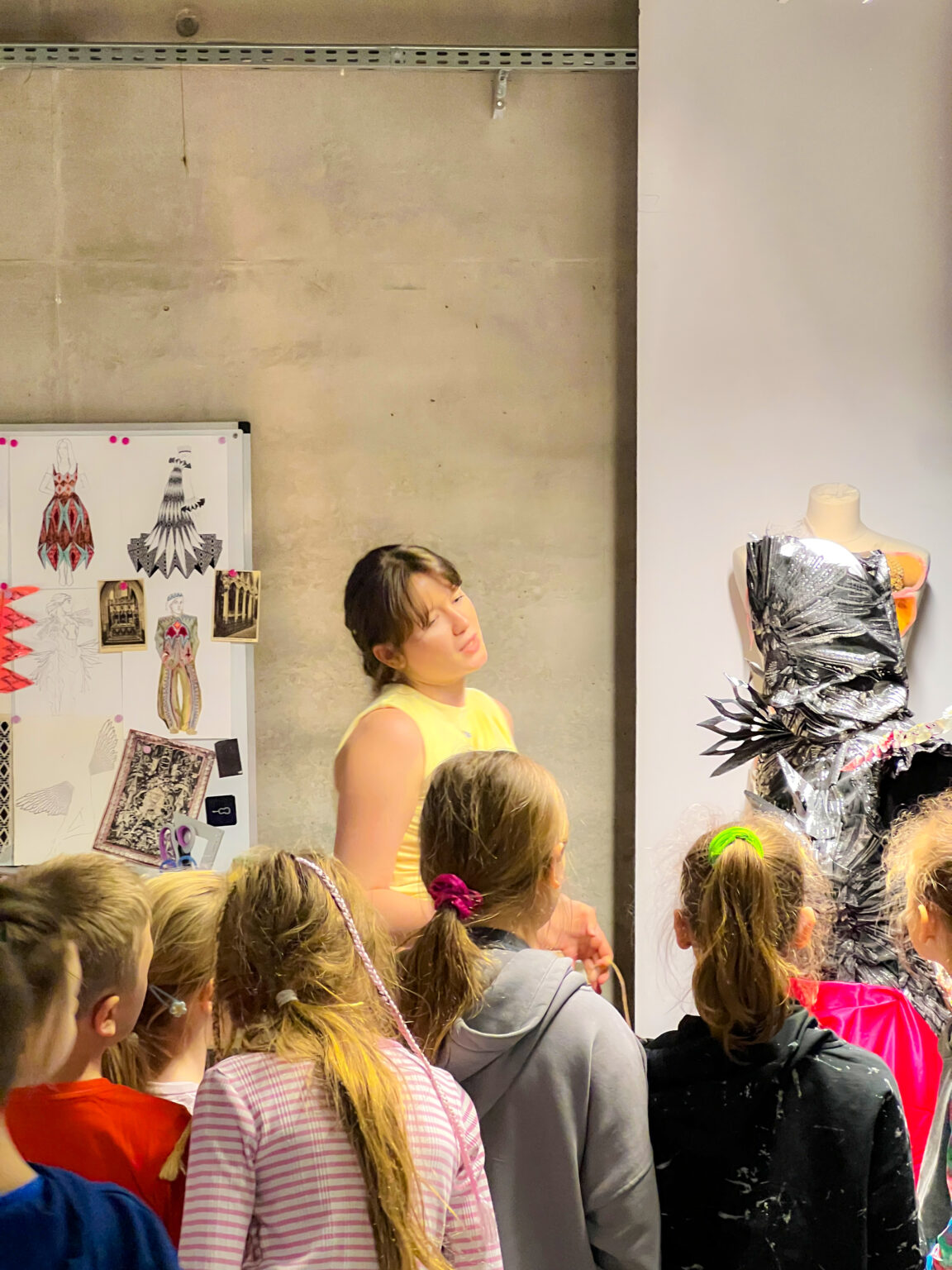 Grupa dzieci stojących tyłem przygląda się dorosłej mówiącej kobiecie. Po prawej widać tułów manekina bez rąk i głowy z udrapowaną na nim srebrną formą kostiumu. Po lewej stronie na tablicy wiszą kolorowe i czarno-białe szkice postaci w kostiumach i monochromatyczne zdjęcia.