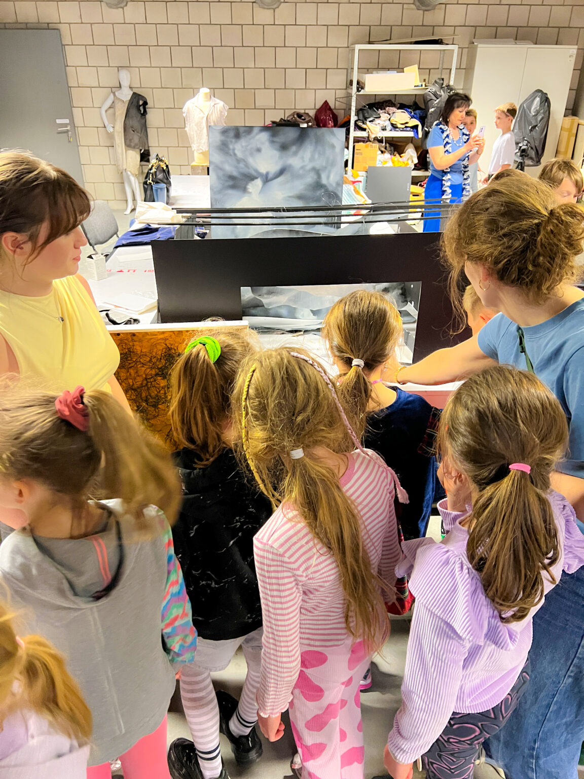 Grupa dzieci stojących tyłem ogląda pudło makiety sceny. Po prawej i lewej stronie stoją dorosłe kobiety. W tle widać manekiny i otwartą szafę z chaotycznie poukładanymi pudłami i szmatami.