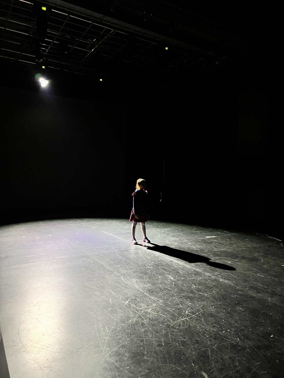 Młoda dziewczynka stoi po środku ciemnego pomieszczenia oświetlona mocnym białym światlem reflektora od tyłu.