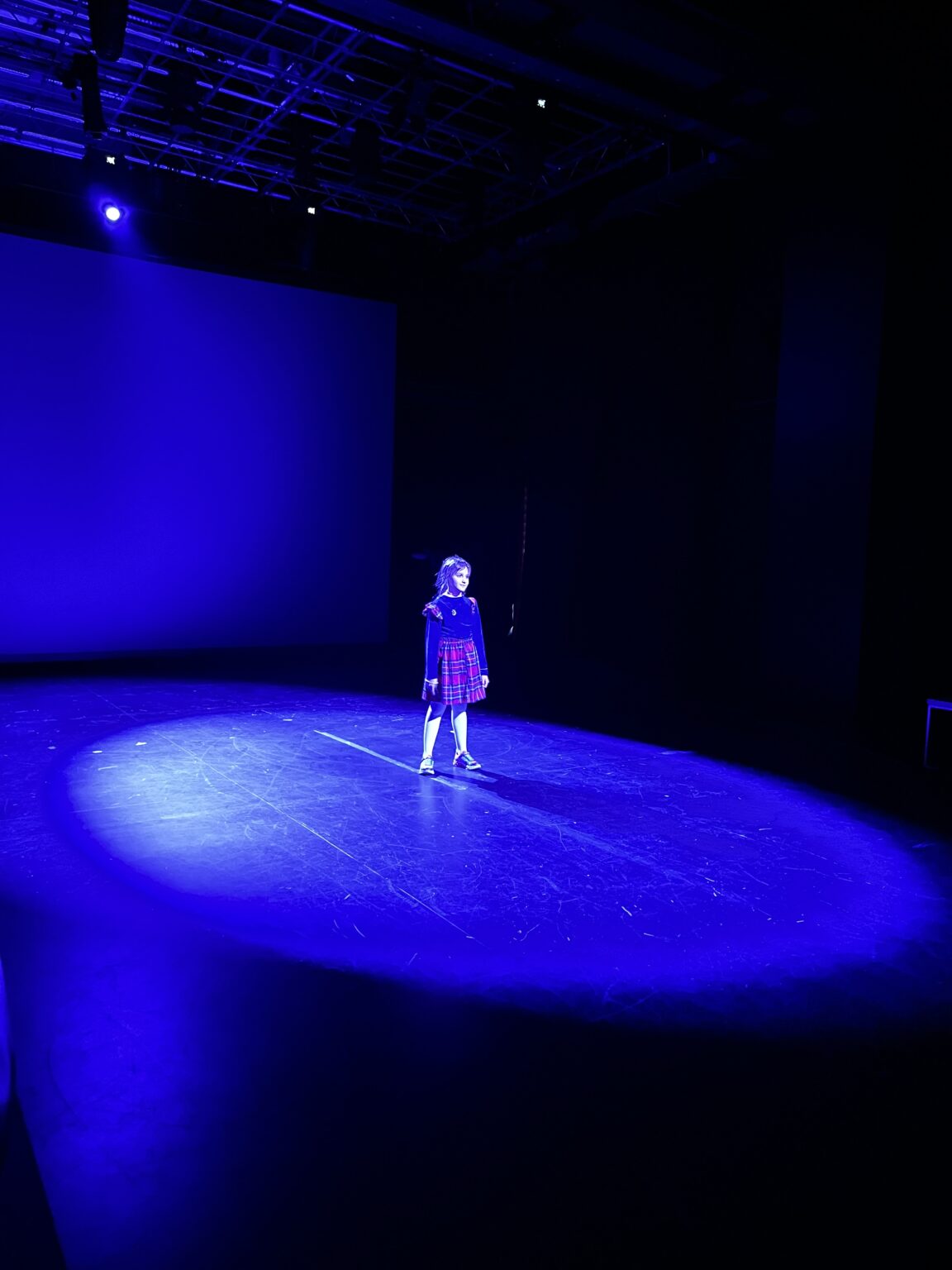 Młoda dziewczynka stoi po środku ciemnego pomieszczenia oświetlona mocnym niebieskim światlem reflektora od tyłu oraz od przodu.