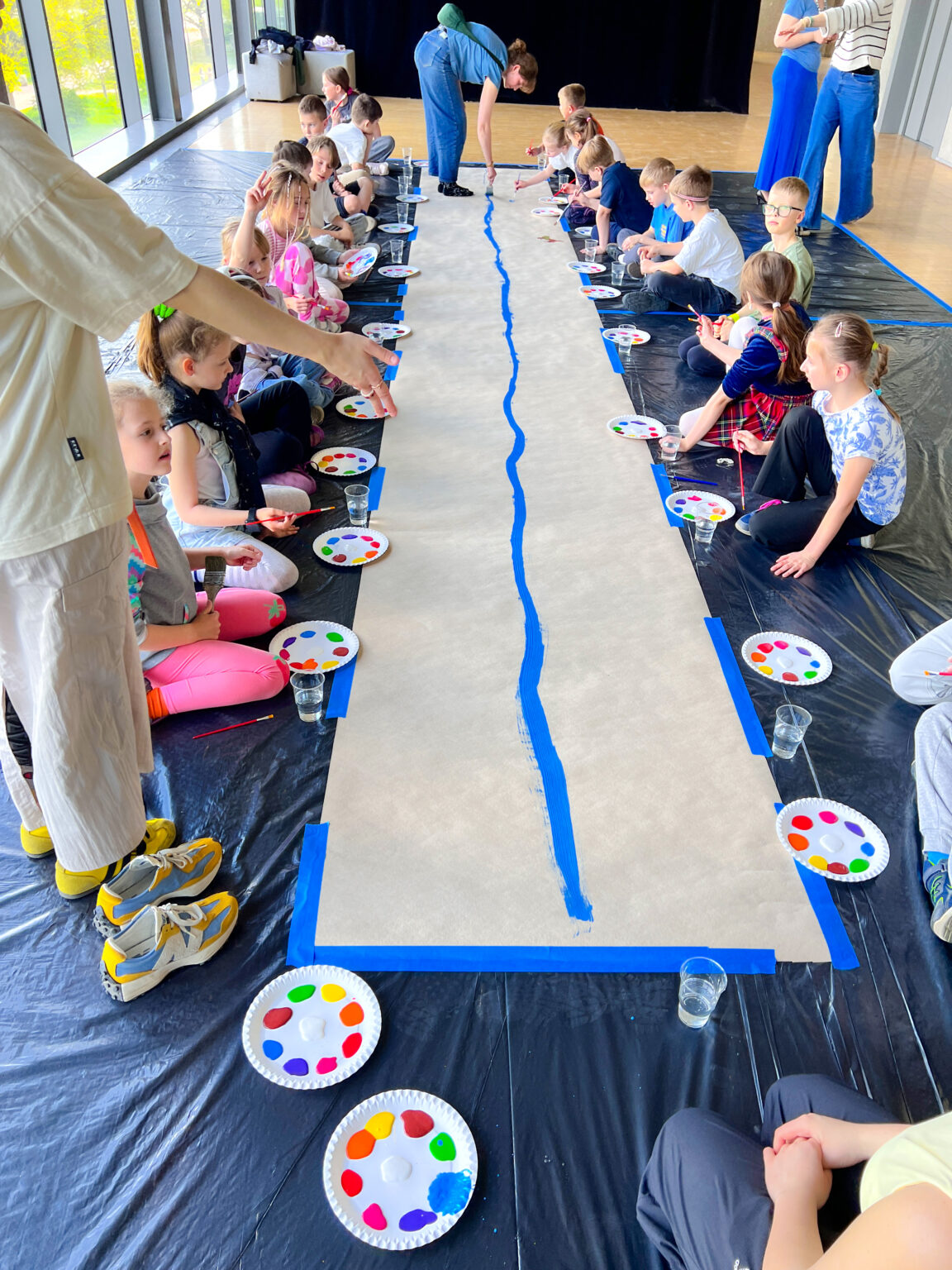 Grupa dzieci siedzi na pokrytej czarną folią podłodze. Obok siebie leżą tekturowe talerze z plamami kolorowych farb. Po środku znajduje się długi pas szaro-brązowego papieru obklejonego niebieską taśmą dookoła. Po środku papieru znajduje się nierówna niebieska linia, którą maluje za pomocą pędzla młoda kobieta na końcu papieru.