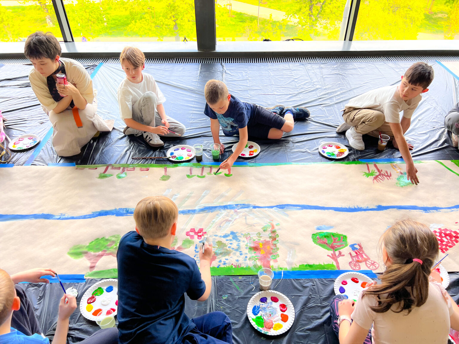 Grupa dzieci widzianych z góry siedzi na pokrytej czarną folią podłodze. Obok leżą tekturowe talerze z plamami kolorowych farb. Po środku znajduje się długi pas szaro-brązowego papieru obklejonego niebieską taśmą dookoła. Po środku papieru znajduje się nierówna niebieska linia a powyżej i poniżej linii powstają malowane przez dzieci drzewa i budynki i inne kolorowe nieokreślone formy.