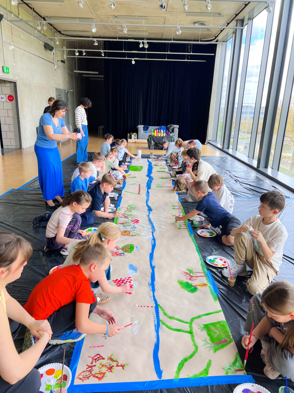 Grupa dzieci widzianych siedzi na pokrytej czarną folią podłodze. Obok leżą tekturowe talerze z plamami kolorowych farb. Po środku znajduje się długi pas szaro-brązowego papieru obklejonego niebieską taśmą dookoła. Po środku papieru znajduje się nierówna niebieska linia a prawej i lewej stronie tej linii powstają malowane przez dzieci drzewa i budynki i inne kolorowe nieokreślone formy.