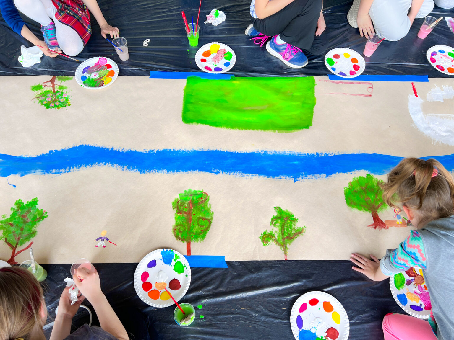 Grupa dzieci widzianych z góry siedzi na pokrytej czarną folią podłodze. Obok leżą tekturowe talerze z plamami kolorowych farb. Po środku znajduje się długi pas szaro-brązowego papieru obklejonego niebieską taśmą dookoła. Po środku papieru znajduje się nierówna niebieska linia a powyżej i poniżej linii powstają malowane przez dzieci drzewa i budynki i inne kolorowe nieokreślone formy.