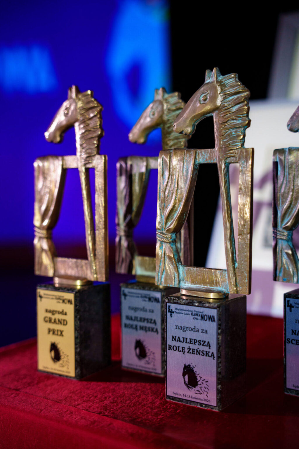 Grupa stojących na czerwonym welurze nagród. Odlane metalowe figury przedstawiają głowę konia na kiju oraz pustą ramę z zasłoną i są umieszczone na kamiennym cokole z podpisami.