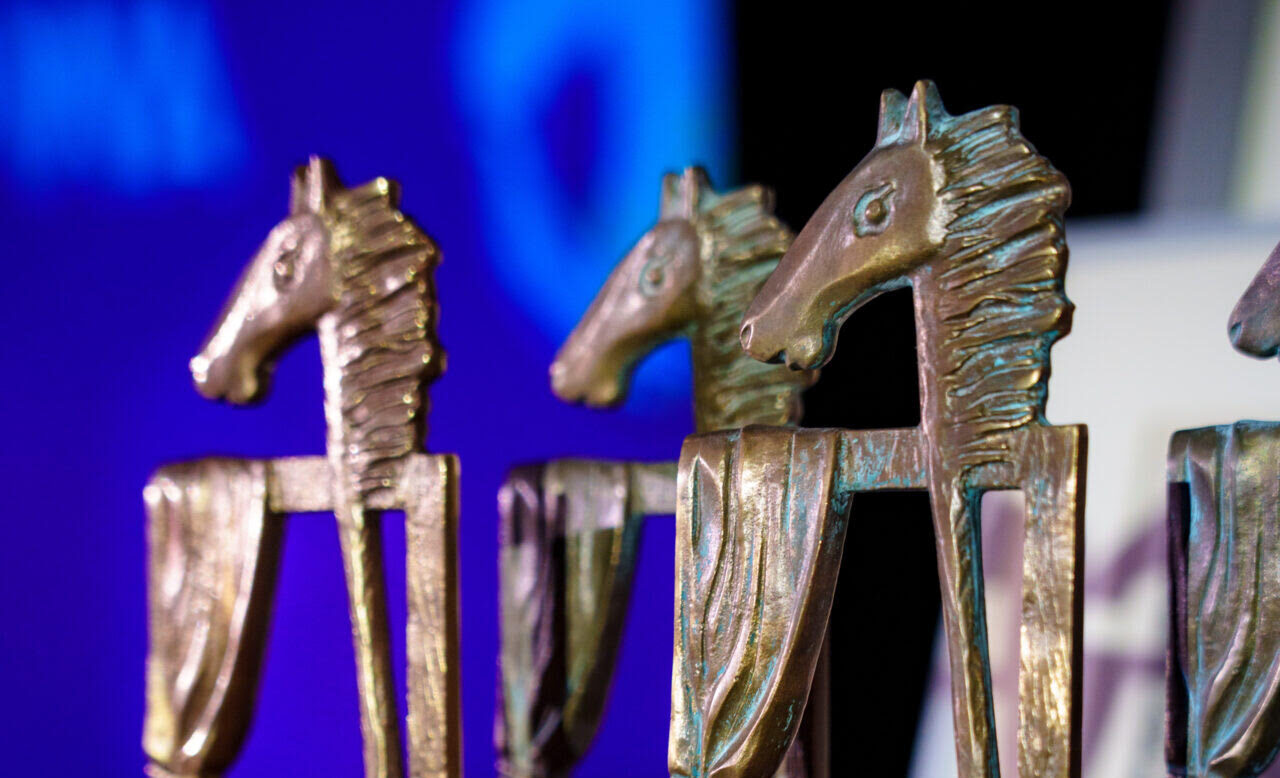 Grupa stojących na czerwonym welurze nagród. Odlane metalowe figury przedstawiają głowę konia na kiju oraz pustą ramę z zasłoną i są umieszczone na kamiennym cokole z podpisami.